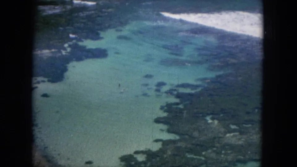 1964 Oahu home-movie frame with turquoise reef shallows, dark rock or coral shapes, and white waves breaking near shore.