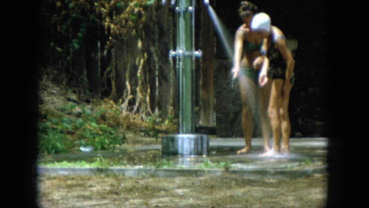 At the outdoor shower, the swim-capped woman rinses her lower legs while her companion stands beside the metal posts.