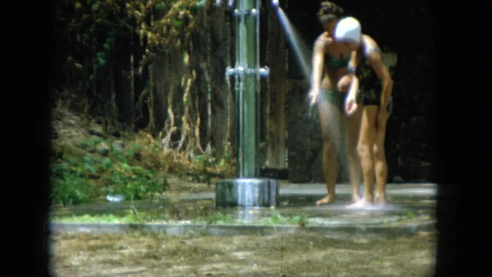 At the outdoor shower, the swim-capped woman rinses her lower legs while her companion stands beside the metal posts.