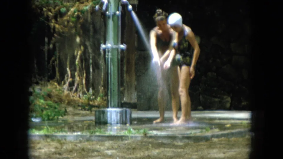 Two women in 1960s-style swimwear stand close at a metal outdoor shower, framed by foliage and a low stone wall.
