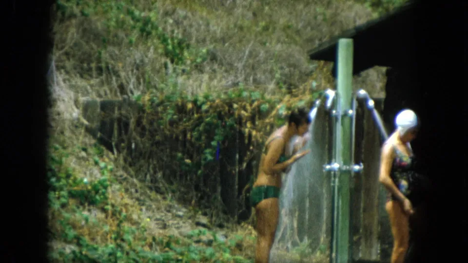 The women separate at the outdoor shower, with one rinsing near the spray and the swim-capped woman walking out of frame.