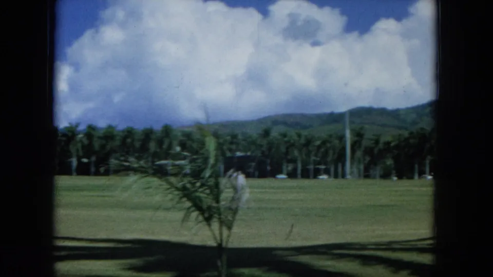 1964 Oahu 8mm frame showing a wide grass field, palm trees, low hills, and clouds viewed through dark framing.