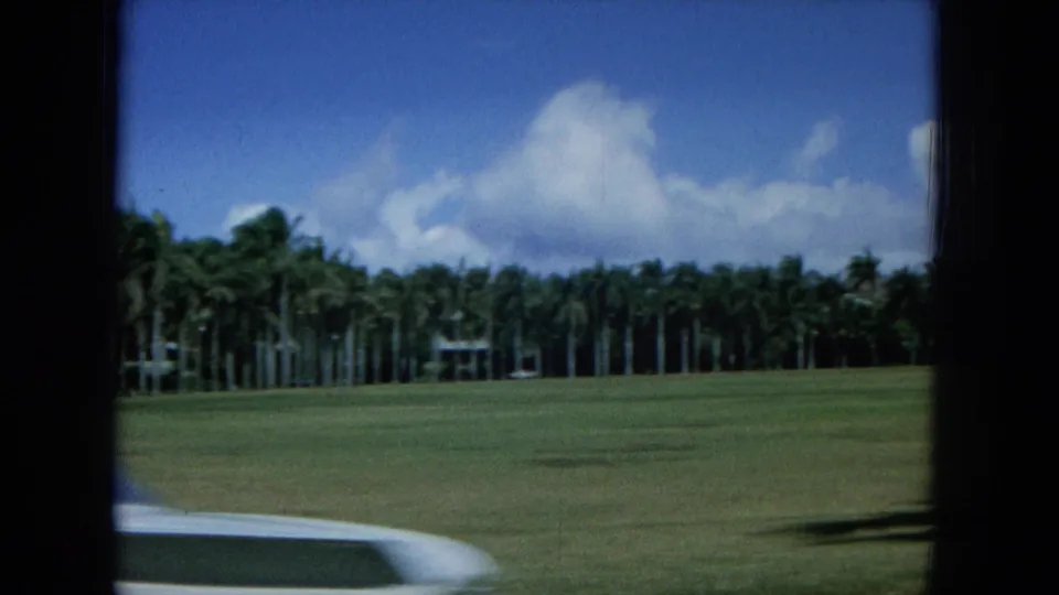 1964 Oahu home-movie frame with palms beyond a grass field, clouds overhead, and a pale curved edge entering the foreground.