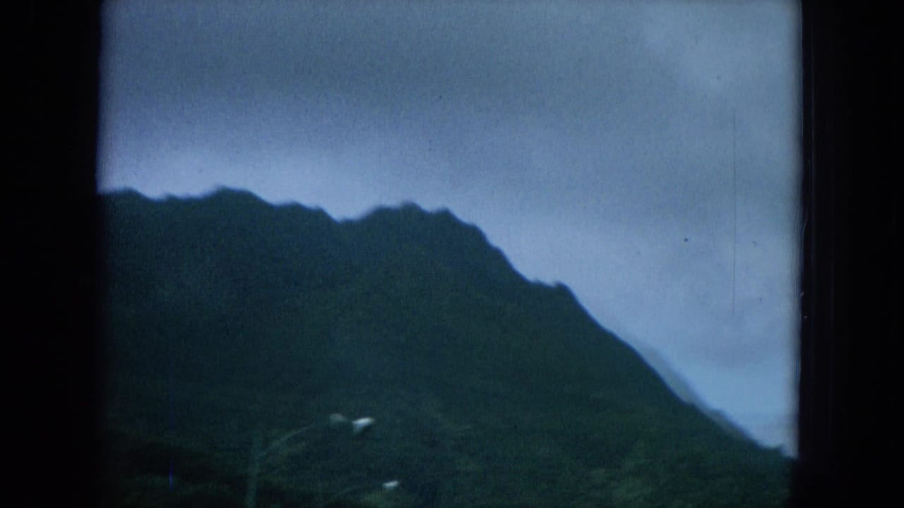 1964 Hawaii home-movie frame showing a dark green mountain ridge and cloudy sky from a moving travel view.