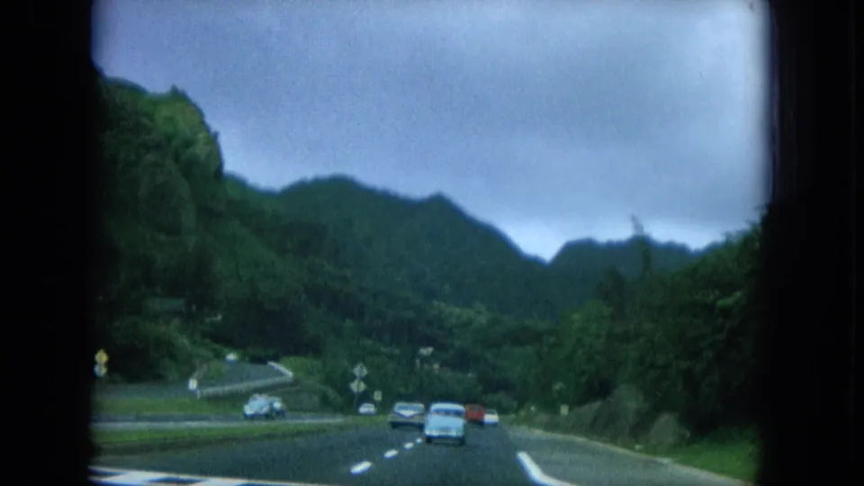 Passenger-view 1964 Hawaii footage of cars on a winding road with wooded slopes and mountain ridges ahead.