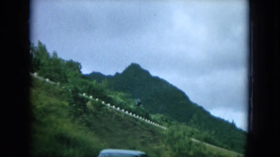 1964 Hawaii home-movie frame showing a roadside hillside, guardrail posts, and layered green mountains from a moving car.