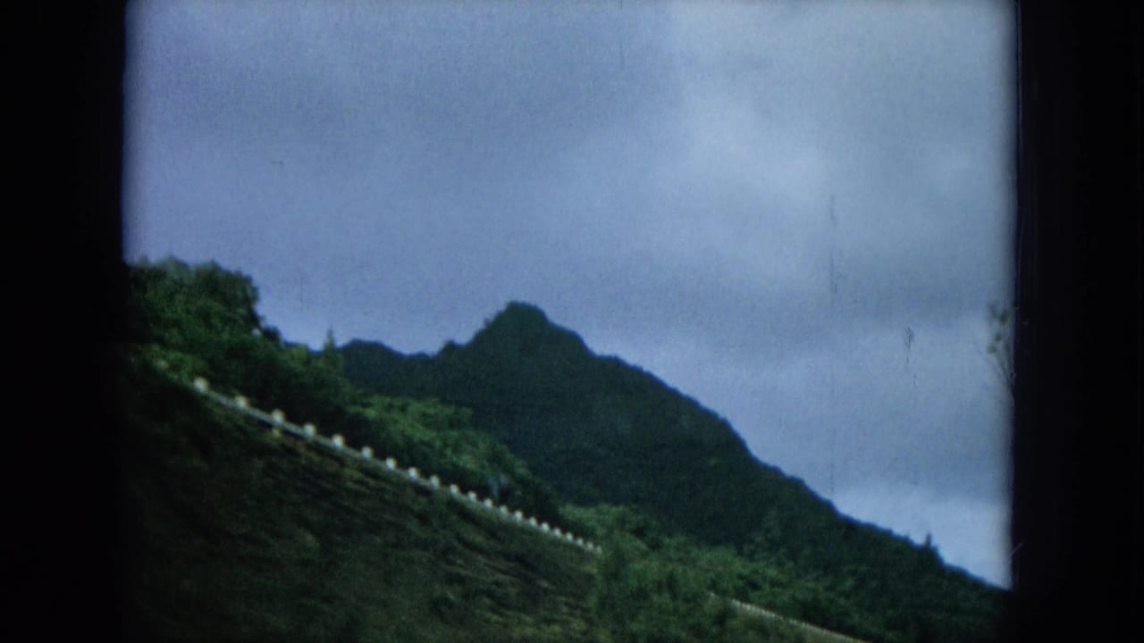Green hillside and distant mountain under a cloudy sky, seen from a moving roadside view.
