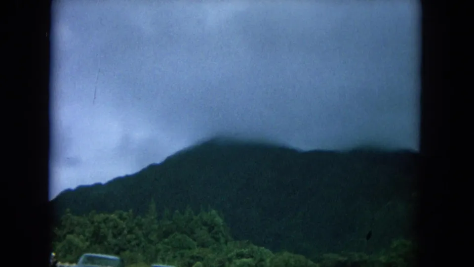 1964 8mm Oahu road view showing a clouded mountain ridge, roadside greenery, and the top of a passing vehicle.
