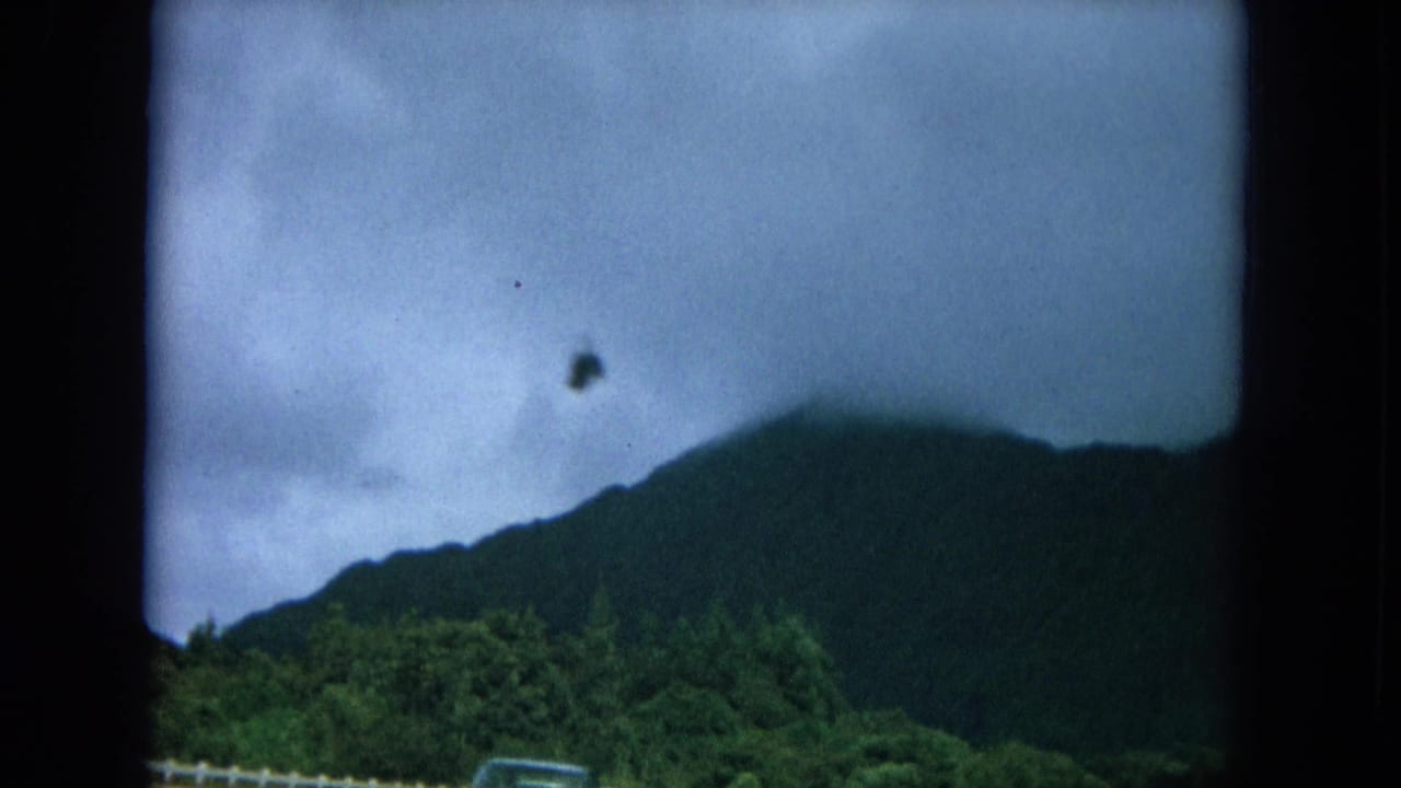 1964 Hawaii home-movie frame showing a dark mountain ridge, cloudy sky, and green roadside vegetation from a moving vehicle.