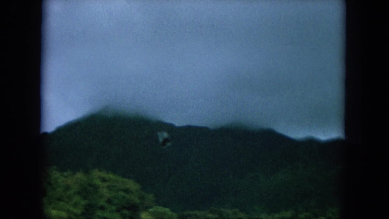 1964 Hawaii frame with a misty green mountain ridge under cloud cover, filmed with soft focus and visible frame edges.