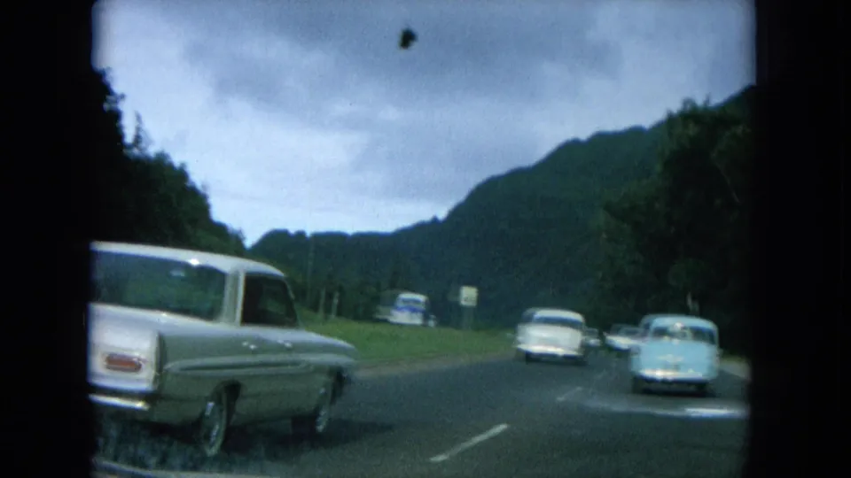 1964 Hawaii road scene with mid-century cars traveling along a paved roadway bordered by trees and mountain slopes.