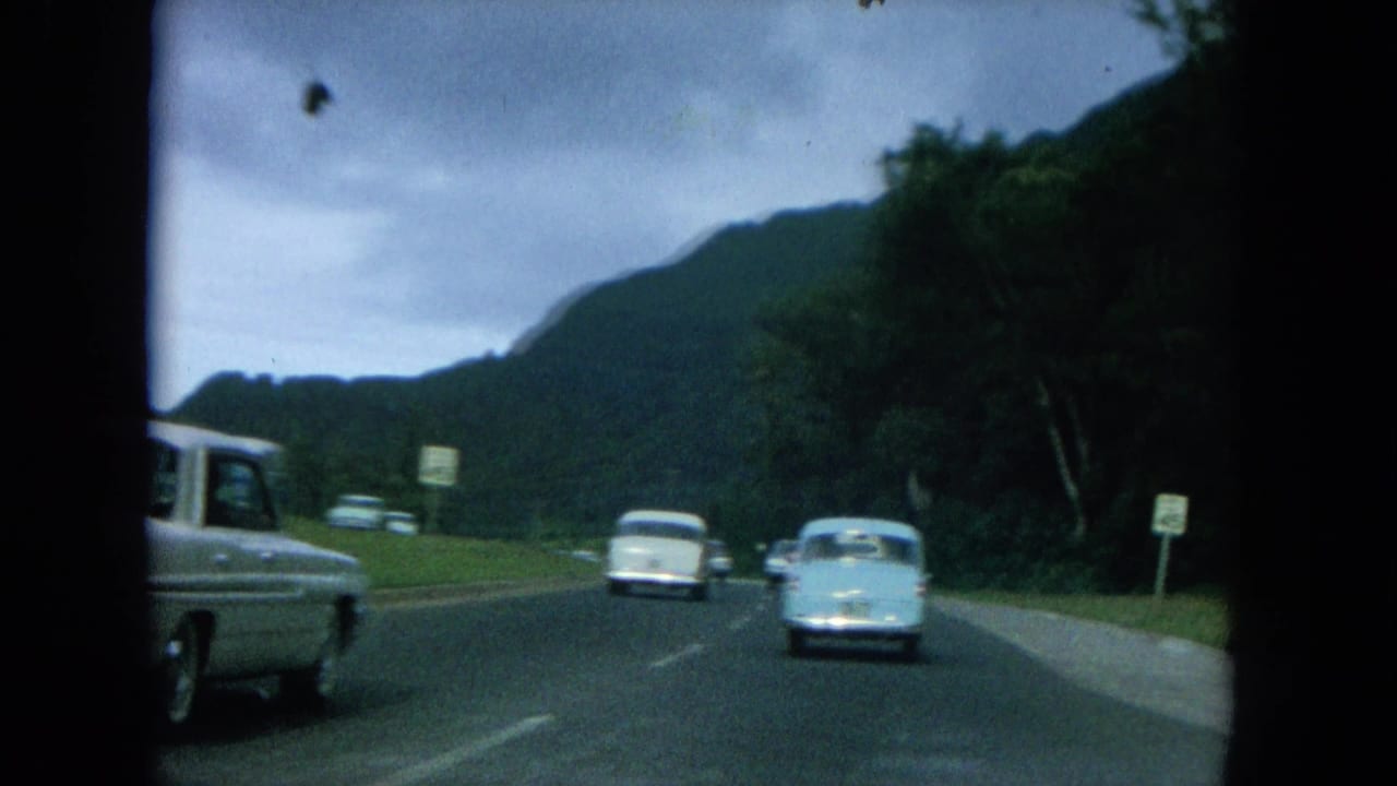 1964 Oahi, Hawaii home-movie frame showing classic cars on a two-lane mountain road under a cloudy sky.