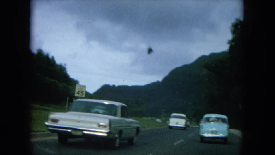 1964 Oahi, Hawaii road scene with a light-colored sedan, distant classic cars, green hills, and a 45 mph sign.