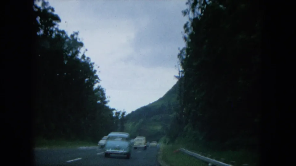 1964 Oahi, Hawaii home-movie view of classic cars moving away on a curved road through dense roadside foliage.