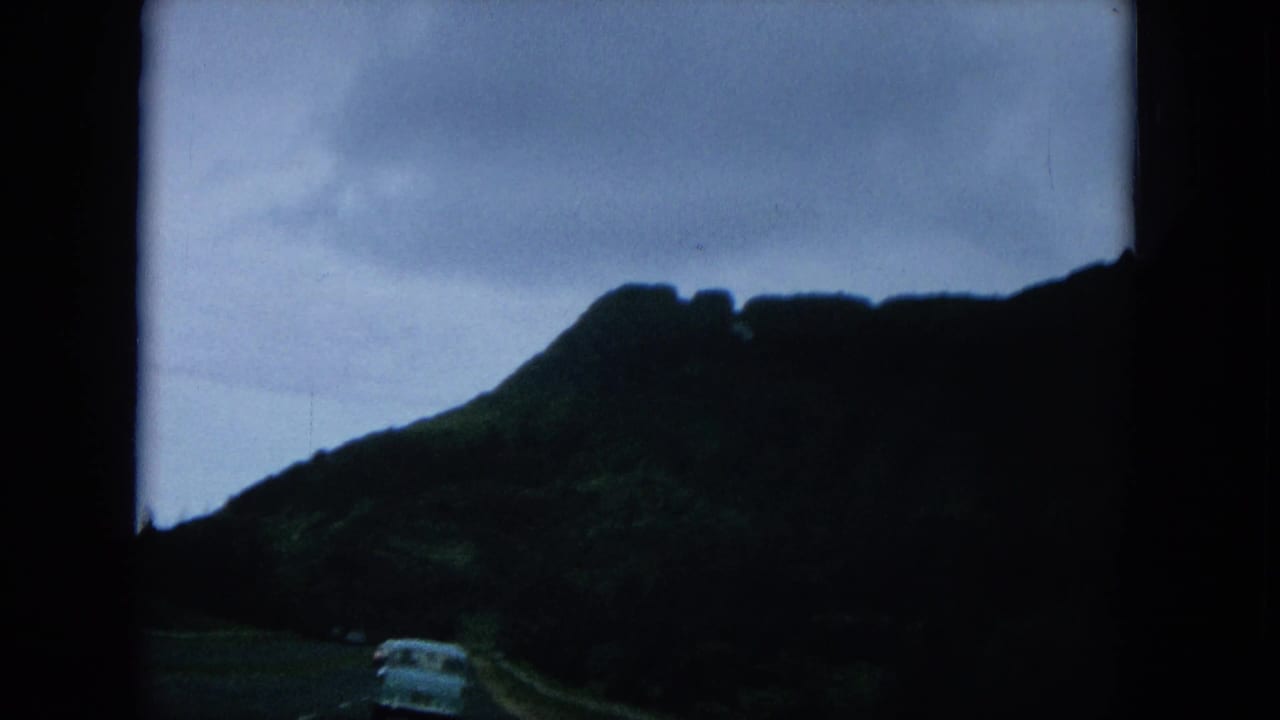Car ahead on a road below dark mountain ridges and cloudy sky.