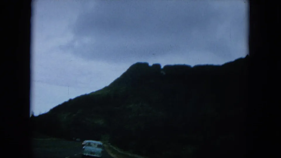 1964 Hawaii travel footage shows a car ahead on a mountain road, with steep ridges silhouetted against an overcast sky.