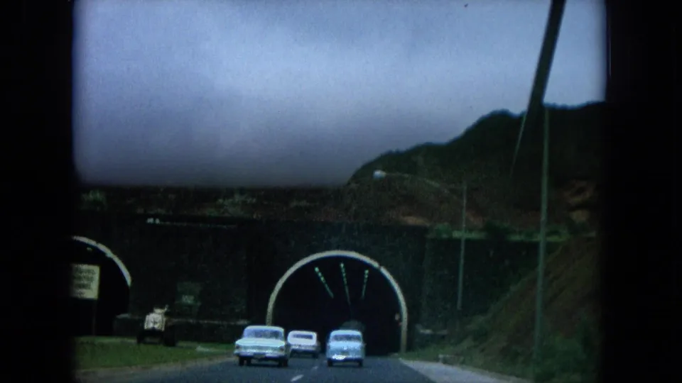 1964 Hawaii 8mm frame showing cars approaching twin tunnel openings on a mountain road, with ridges and a cloudy sky behind them.