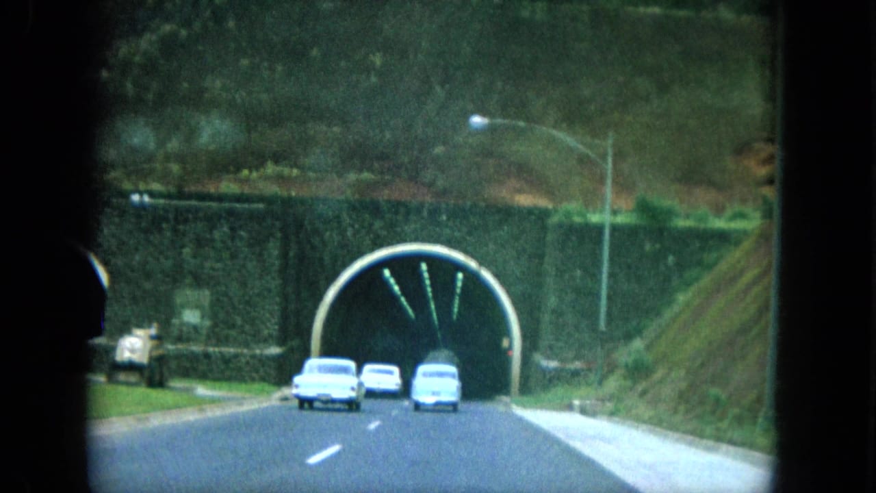 1964 8mm home-movie view on Oahu showing several cars driving toward a stone-arched tunnel beside green hills.
