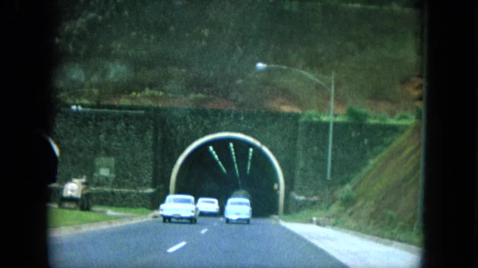 1964 8mm home-movie view on Oahu showing several cars driving toward a stone-arched tunnel beside green hills.