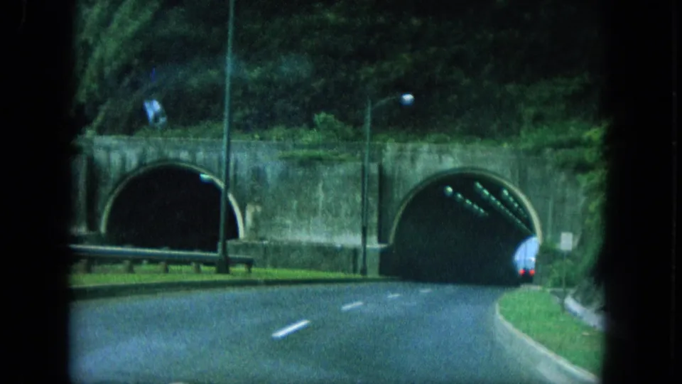 The moving-camera view follows a road curving toward tunnel portals set into a concrete or stone wall on Oahu.