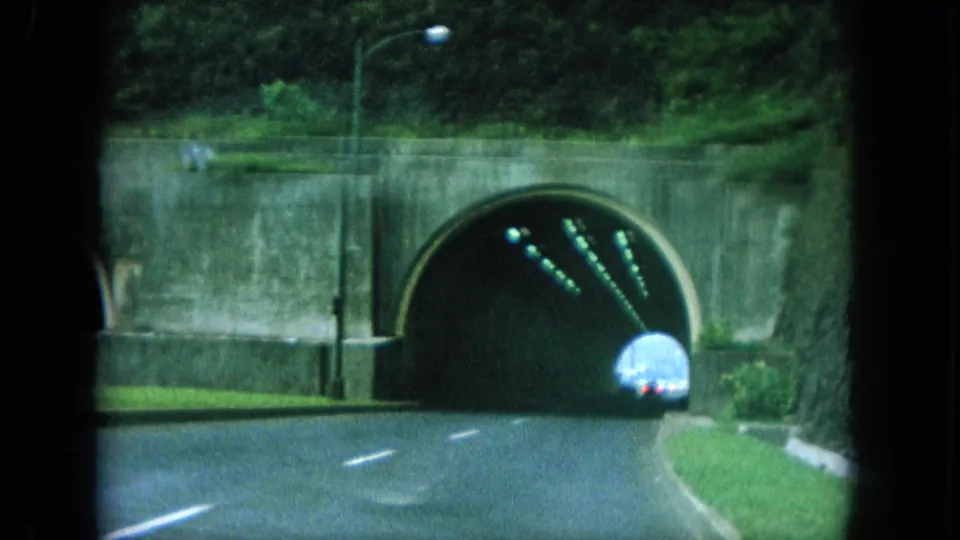A vehicle enters the dark tunnel as the 8mm camera approaches from the road, with green hills and a retaining wall above.