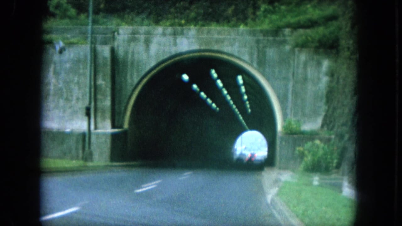 1964 8mm Oahu road-trip frame showing a vehicle approaching a concrete tunnel entrance with lane markings, walls, greenery, and lights inside.