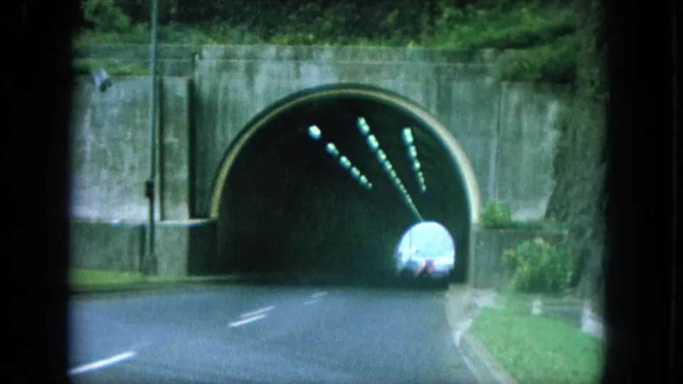 1964 8mm Oahu road-trip frame showing a vehicle approaching a concrete tunnel entrance with lane markings, walls, greenery, and lights inside.