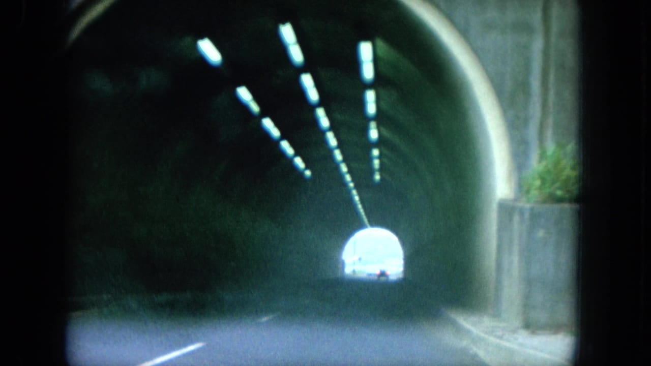 1964 8mm Oahu driving frame looking through a dark concrete tunnel toward a bright exit, with overhead lights receding into the distance.