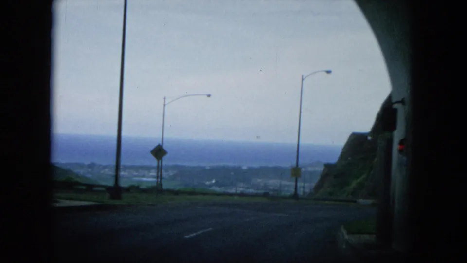 1964 8mm Oahu coastal-road view after a tunnel, showing streetlamps, a cliff edge, roadway, signs, and the ocean horizon in the distance.