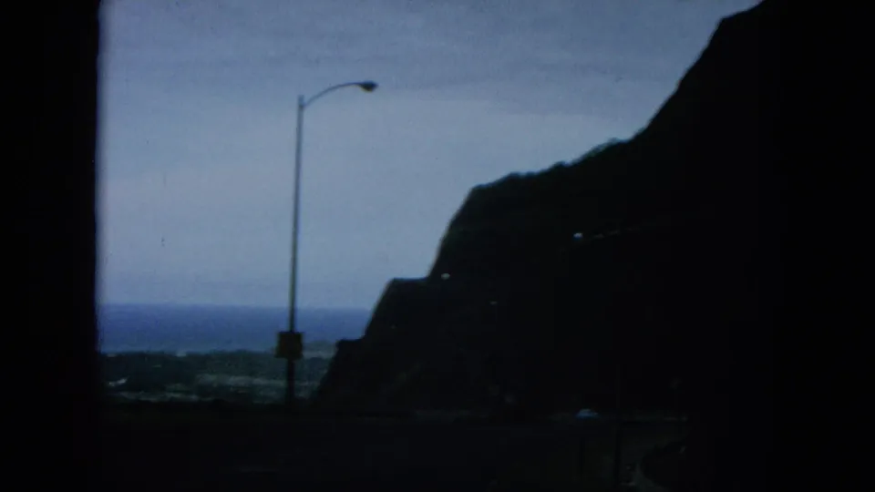 1964 8mm Oahu frame showing a shadowed cliffside road, a single streetlamp, and the ocean horizon beyond the coastal route.