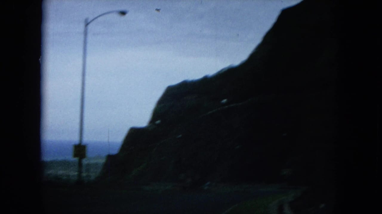 1964 Oahu road view from a moving vehicle, with a silhouetted mountain, ocean horizon, and roadside streetlight.