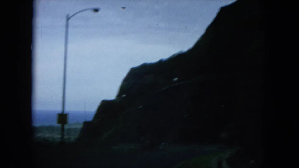 1964 Oahu road view from a moving vehicle, with a silhouetted mountain, ocean horizon, and roadside streetlight.