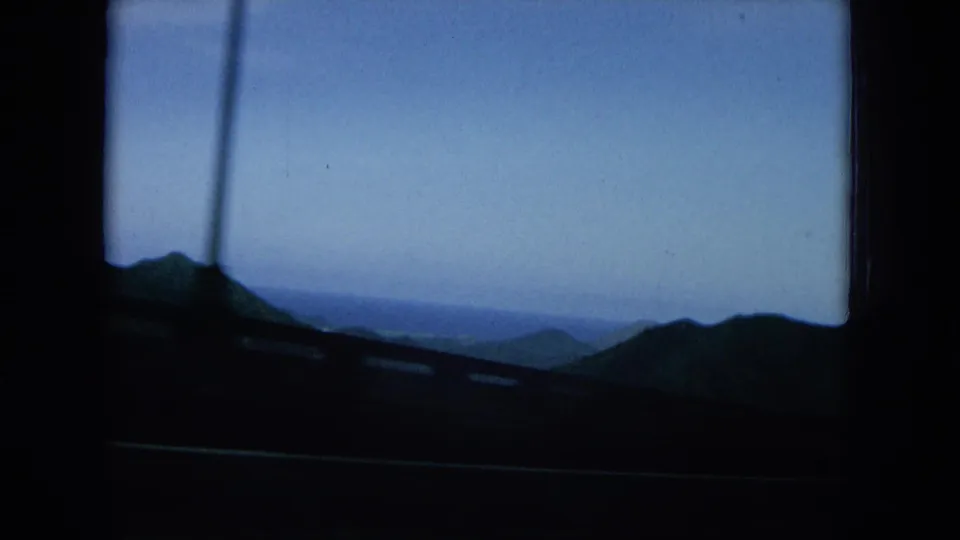 1960s Hawaii home-movie frame looking past a road guardrail toward dark hills, distant water, and a blue sky.