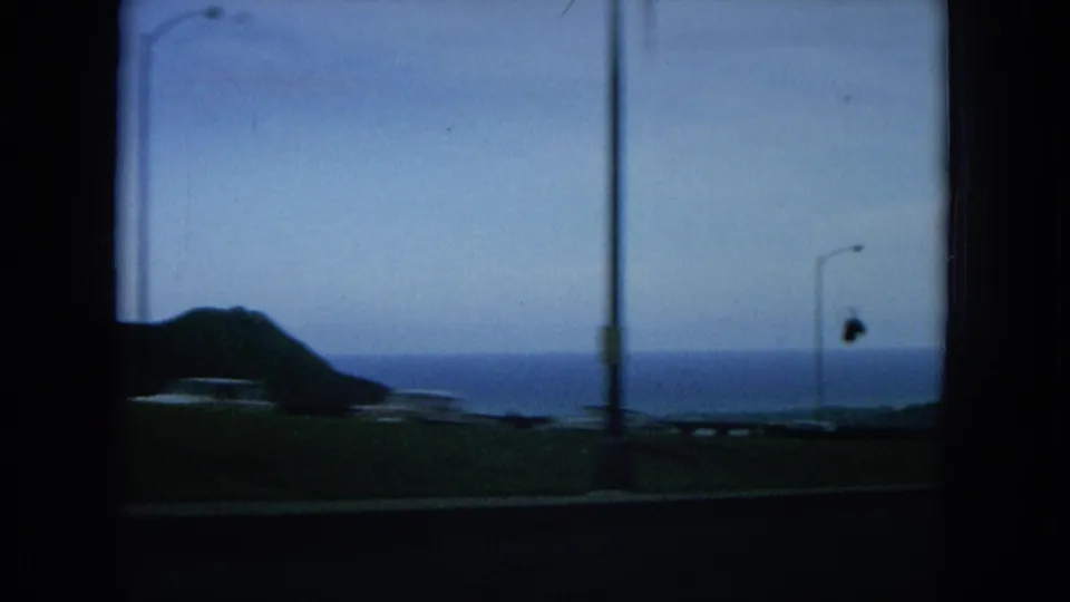 Oahu road descent frame with streetlights, silhouetted hills, coastal water, and dark car-window edges.