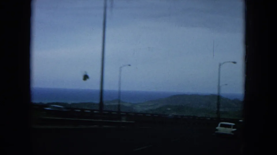 1964 Hawaii home-movie frame showing lamp posts along a coastal road, ocean beyond, distant hills, and a bird overhead.