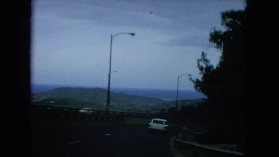 1964 Hawaii 8mm frame of a curving coastal highway with a white car, lamp posts, guardrail, ocean, and hills beyond.