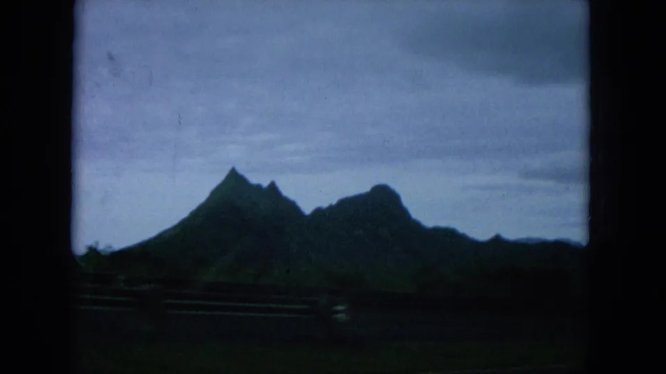 1964 Hawaii home-movie view of a dramatic mountain silhouette beyond roadside railings beneath a cloudy sky.