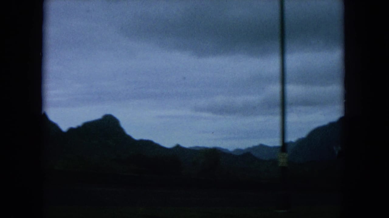 1964 Oahu home-movie frame looking through a car window toward dark mountain ridges under a cloudy sky.