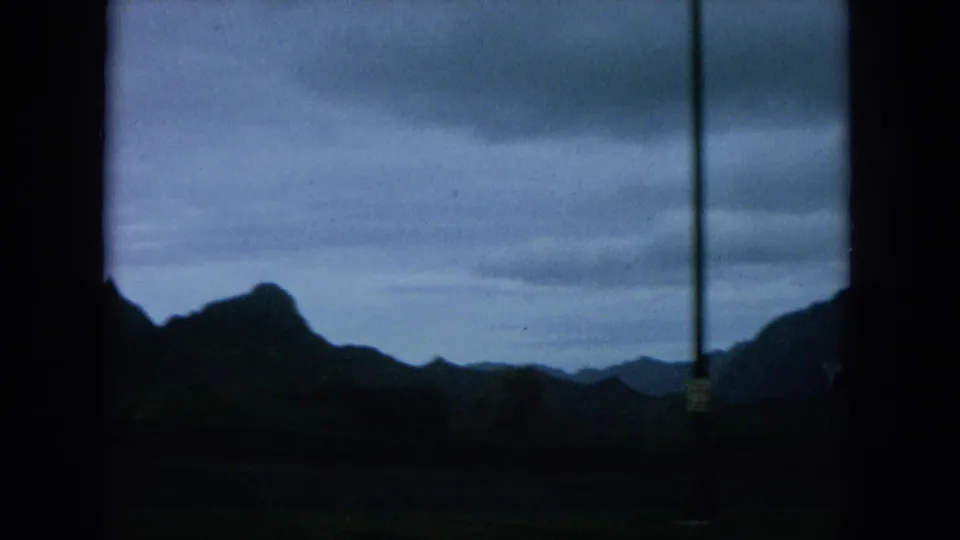 1964 Oahu home-movie frame looking through a car window toward dark mountain ridges under a cloudy sky.