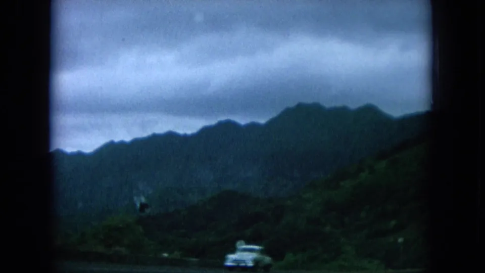 A white car appears on the lower road beneath green Oahu hills and a heavy overcast sky.