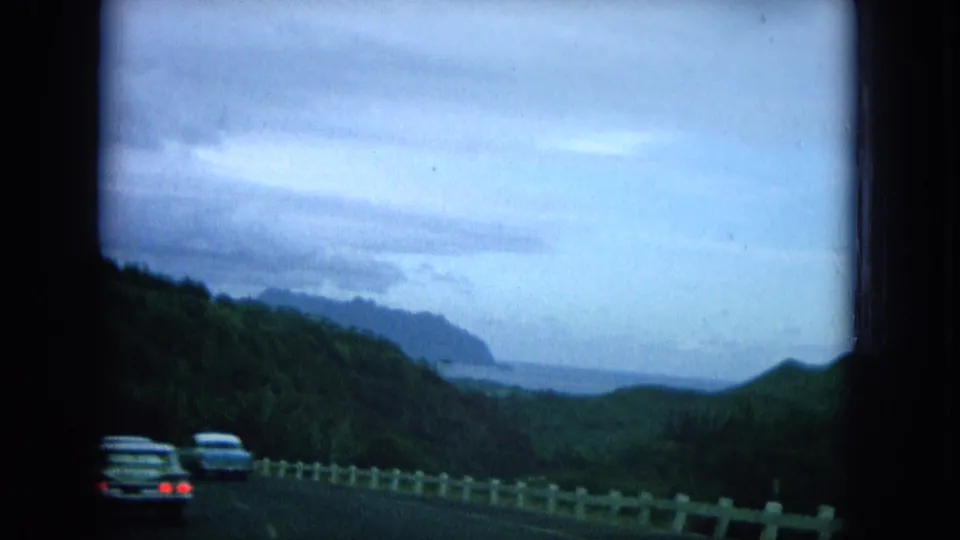 1964 Oahu road scene with cars ahead, a white guardrail, green slopes, and coastline visible in the distance.