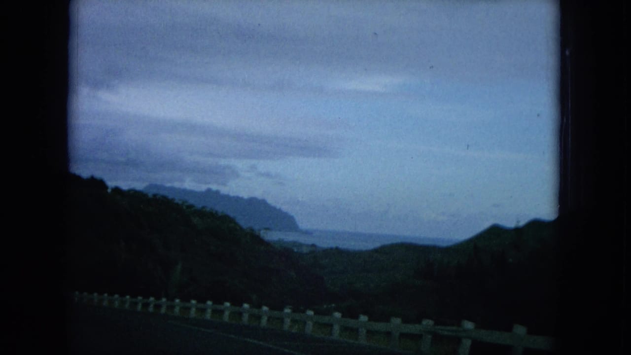 1964 8mm Oahu travel footage showing a winding road, white guardrail, dark green hills, distant ocean, and cloudy sky.