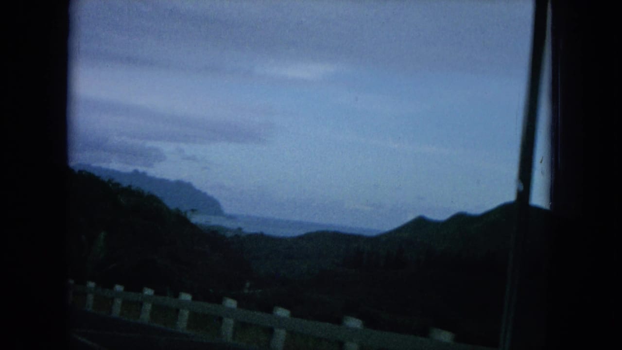 Oahu home-movie frame with a white roadside guardrail, shadowed hills, distant coastline, and overcast sky.