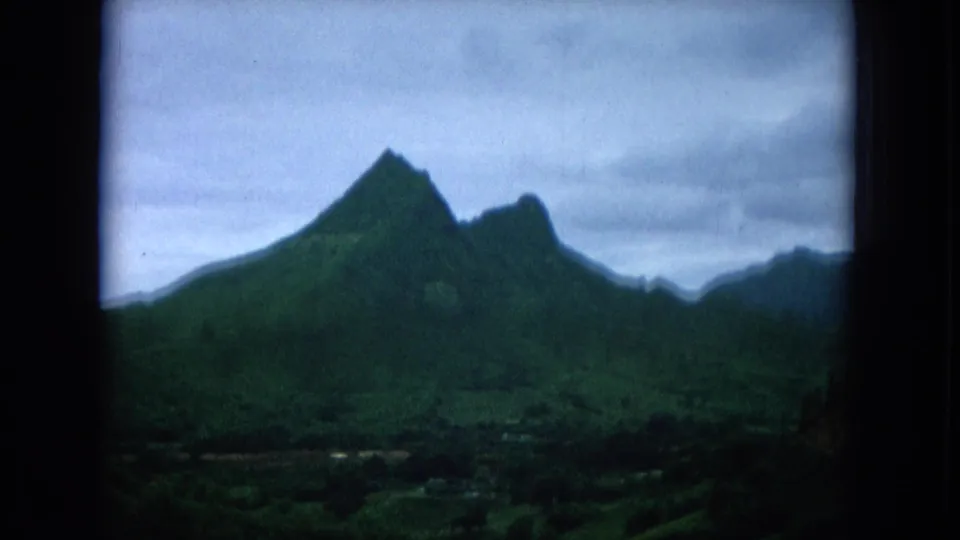 1964 Oahu scenic frame showing sharp green mountain peaks, lower valley vegetation, and a cloudy sky.
