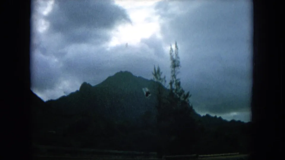 1964 Hawaii roadside view with a dark mountain profile, cloudy sky, and vegetation passing near the lens.