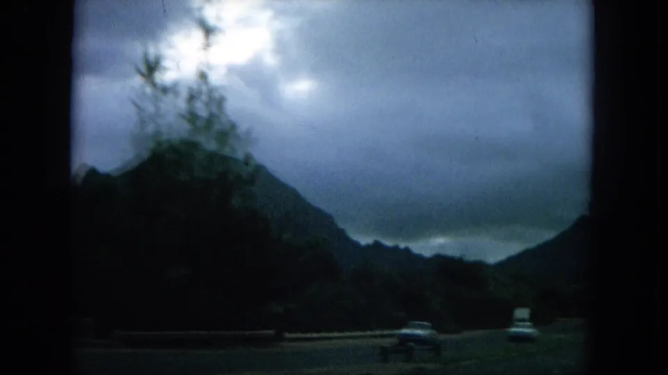 1964 Oahu moving-car footage showing vehicles on a road below dark green mountains and a cloudy sky.