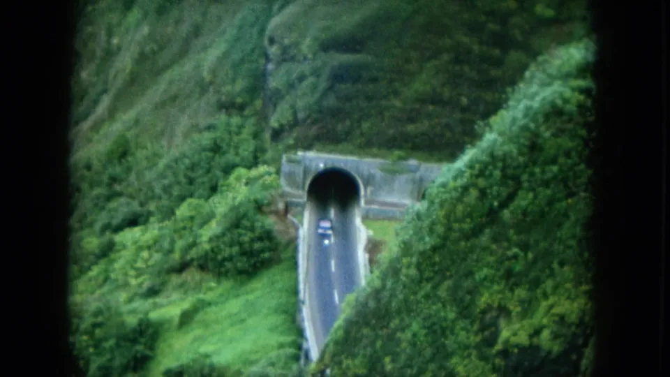 1964 Hawaii home-movie frame of a road and car entering a tunnel cut into a green mountainside.