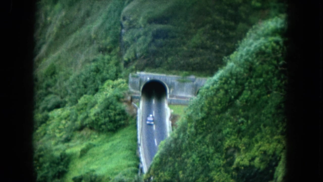 1964 8mm Hawaii travel footage shows a small car on a road entering a tunnel through dense green hillside vegetation.