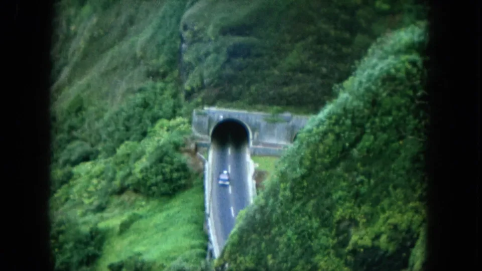 1964 8mm Hawaii travel footage shows a small car on a road entering a tunnel through dense green hillside vegetation.