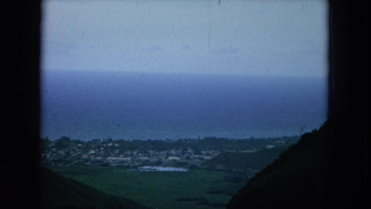 Elevated 1964 Oahu view across dark hills toward a coastal town, fields, and a blue ocean horizon.